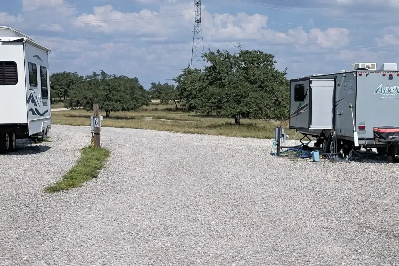 RVs parked on a gravel lot beside a grassy field under a cloudy sky