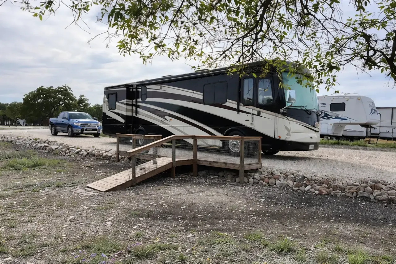 Black and white motorhome parked on gravel beside a wooden ramp, with a blue car and trees nearby.