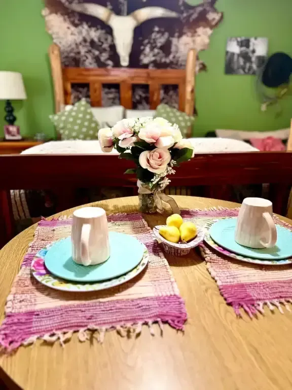 Dining table set for breakfast with cups, plates, flowers, and fruit in a cozy bedroom.