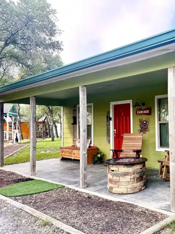 Porch with green siding, red door, wooden bench, and rustic barrel table under a sloped roof.