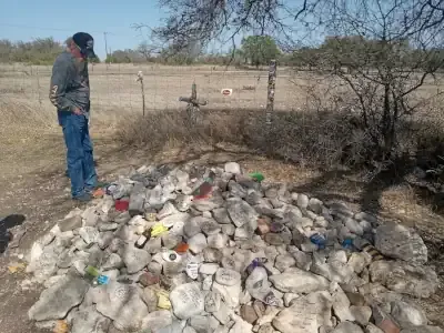 Person standing beside a pile of trash outdoors near a fence and dry brush.