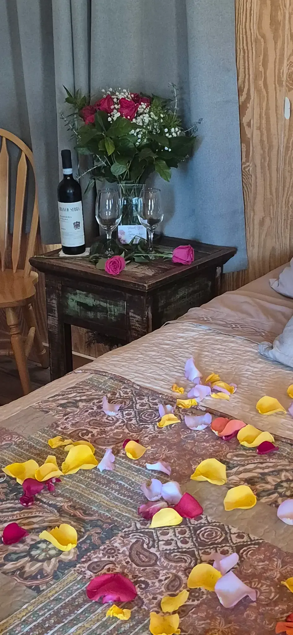 Petals scattered on a bed in a cozy room, with flowers on a bedside table and chair nearby