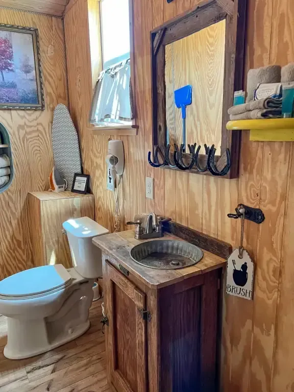 Small rustic bathroom with a toilet, sink, mirror, towel rack, and wood-paneled walls.