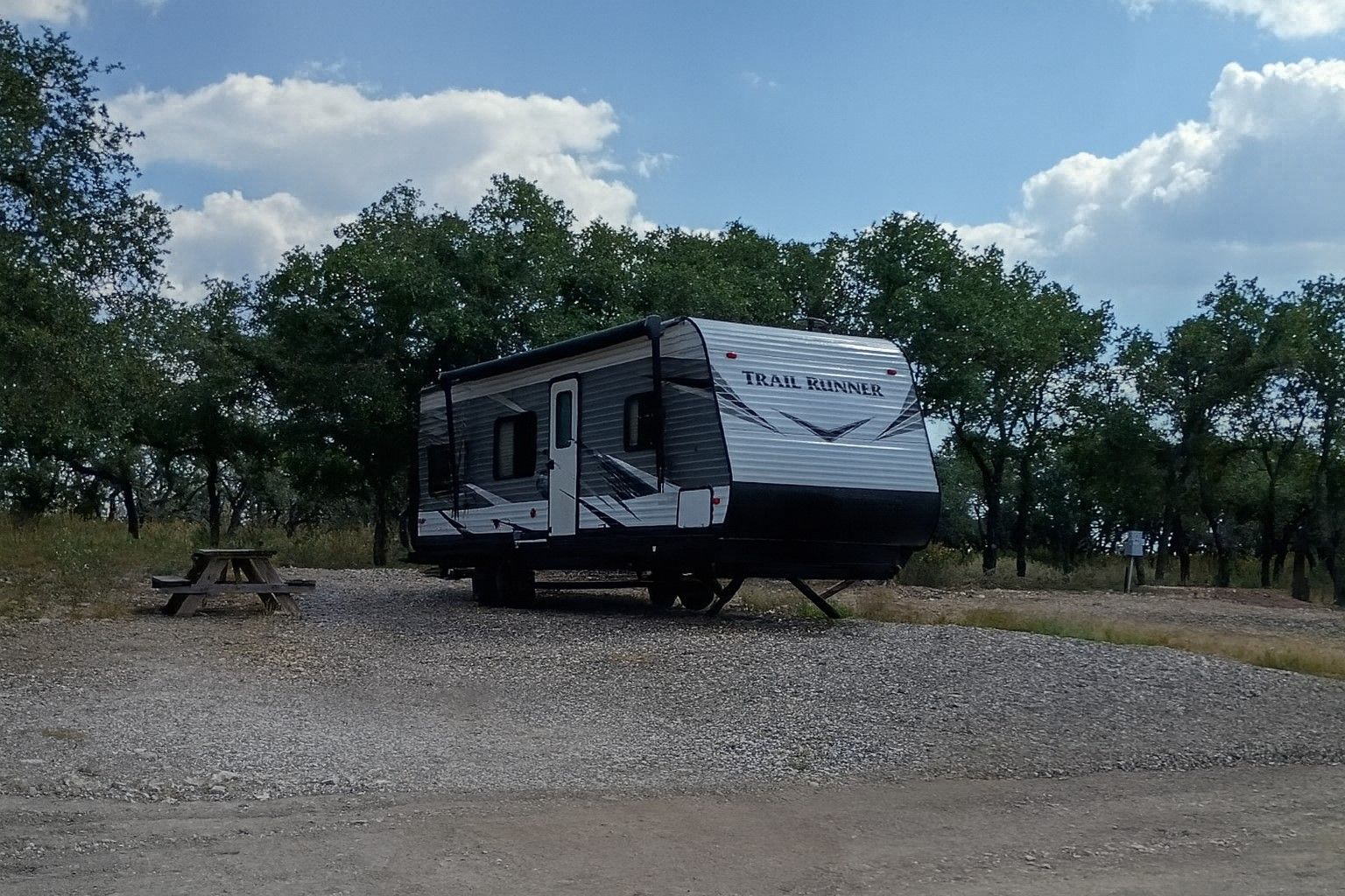 White and black camper trailer parked on gravel under trees and a cloudy sky