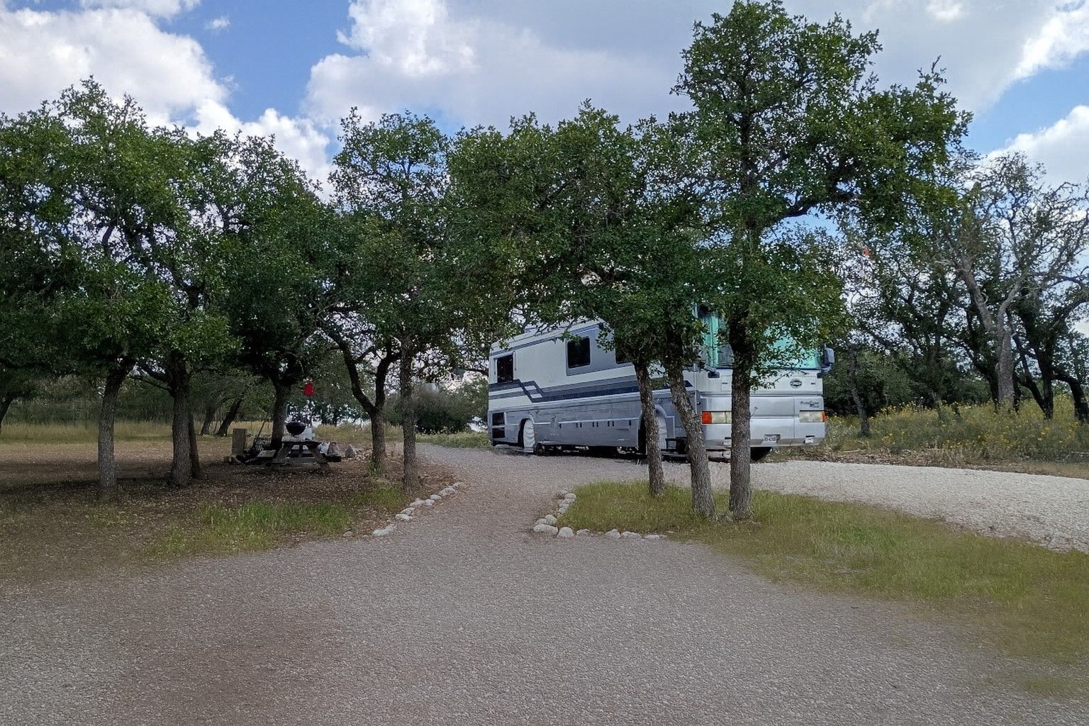 Gravel driveway with a white RV among trees under a partly cloudy sky.