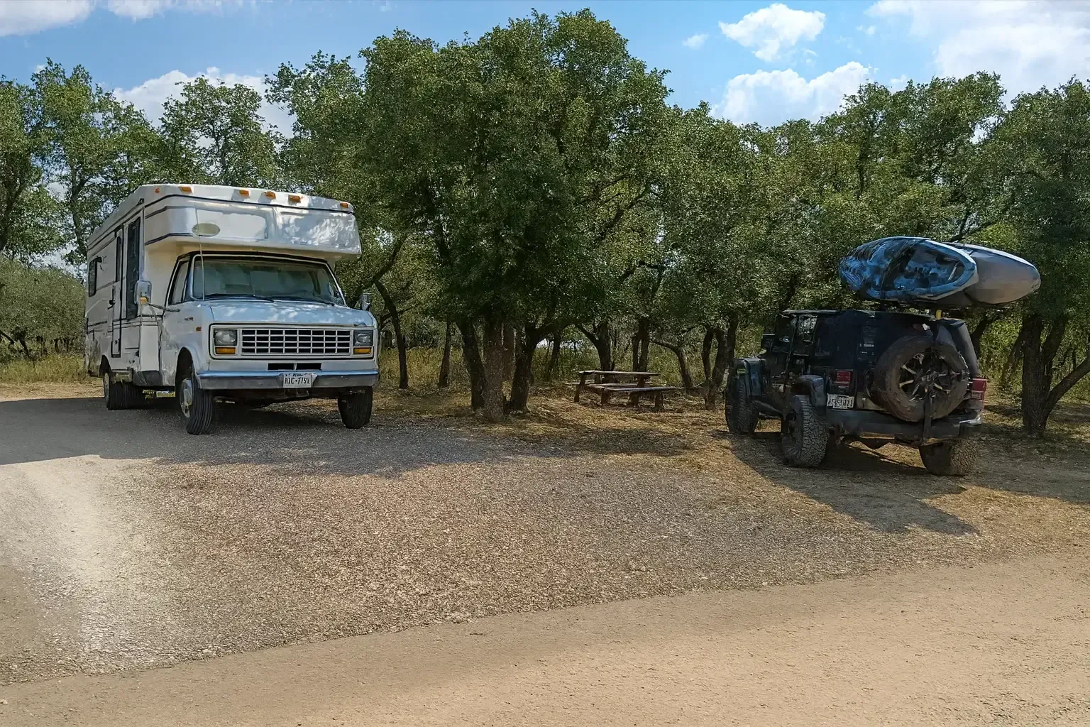 Camper truck and ATV parked on a gravel road beside trees and brush