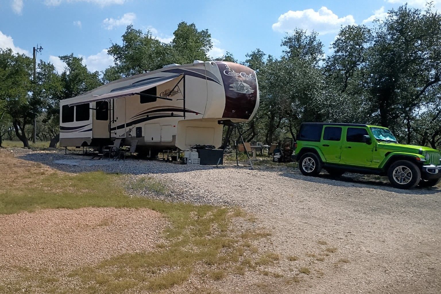 Beige RV and bright green Jeep parked on a gravel lot by trees under a blue sky.