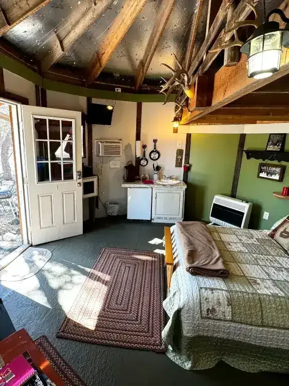 Rustic bedroom with a bed, dresser, rug, open door, and exposed wooden beams in a round room.