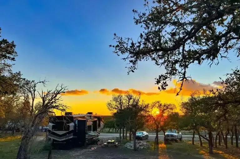 Sunset over a wooded campground with RVs and parked cars beneath silhouetted trees