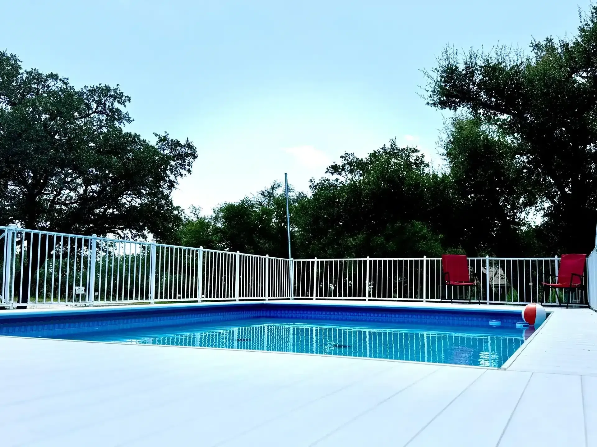 Outdoor swimming pool with blue water, white deck, and red lounge chairs beside trees