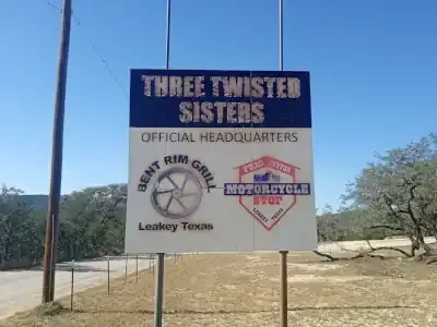 Roadside sign for Three Twisted Sisters official headquarters in Leakey, Texas, under a blue sky.