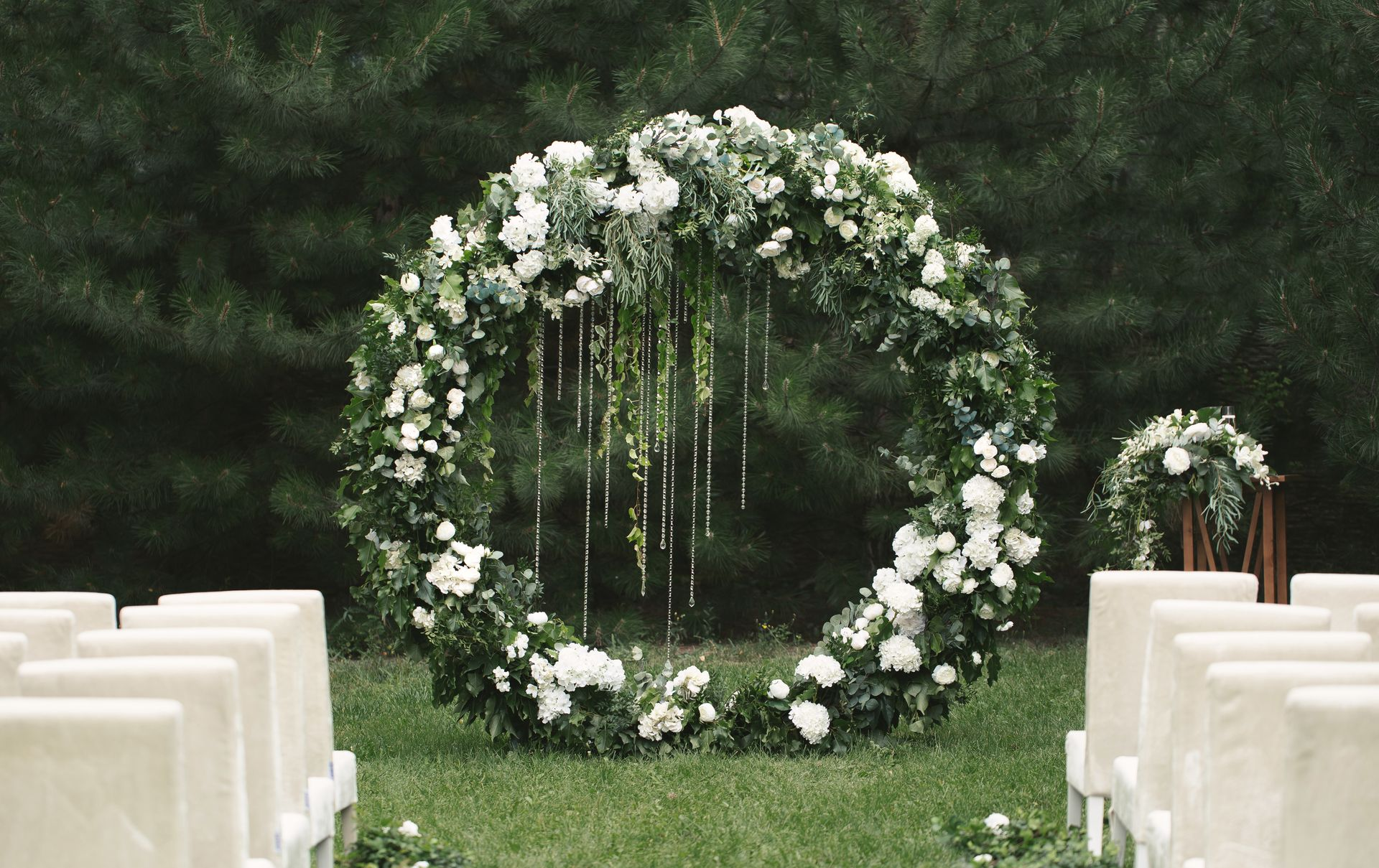 White floral wedding arch on a green lawn, with rows of white chairs on both sides.