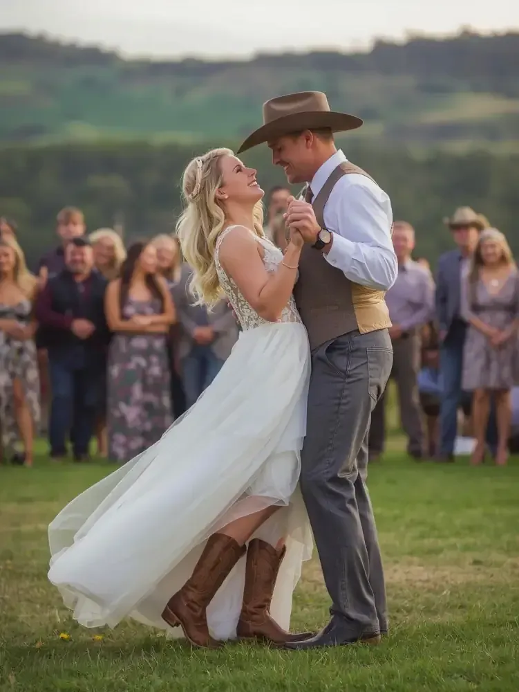 Couple slow dancing outdoors at a rustic wedding, bride in white dress and cowboy boots, groom in hat and vest