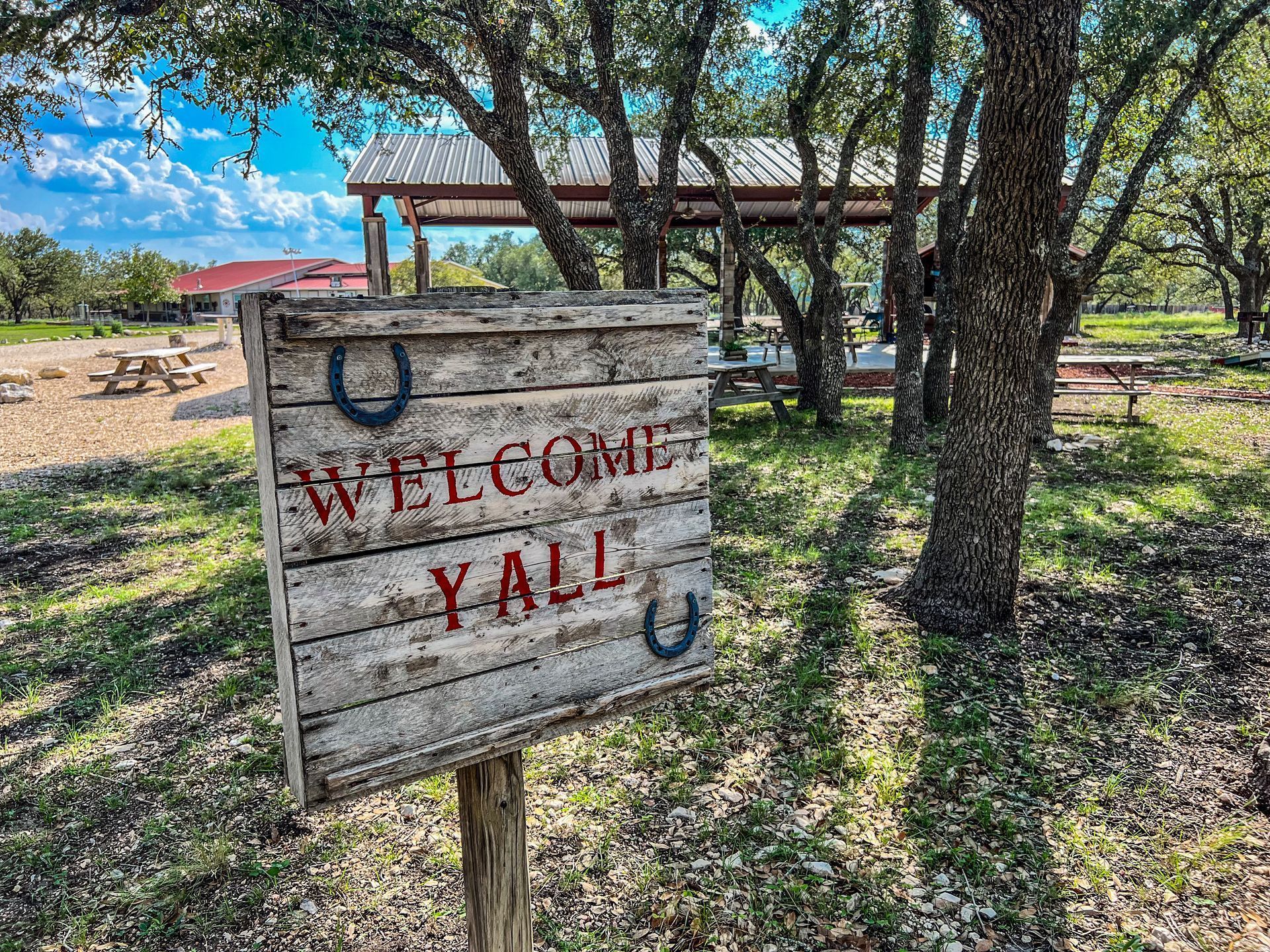 Weathered wooden sign reading “WELCOME YALL” in red, hanging among trees in a rustic outdoor setting