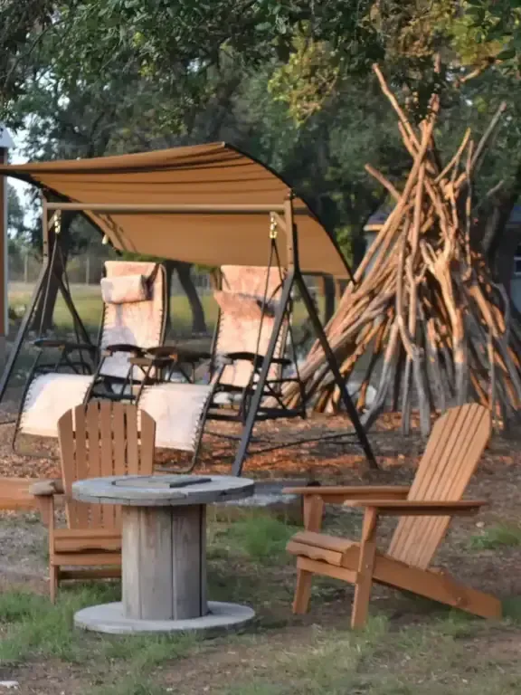 Rustic campsite with canopy chairs, firewood teepee, and wooden chairs around a spool table outdoors