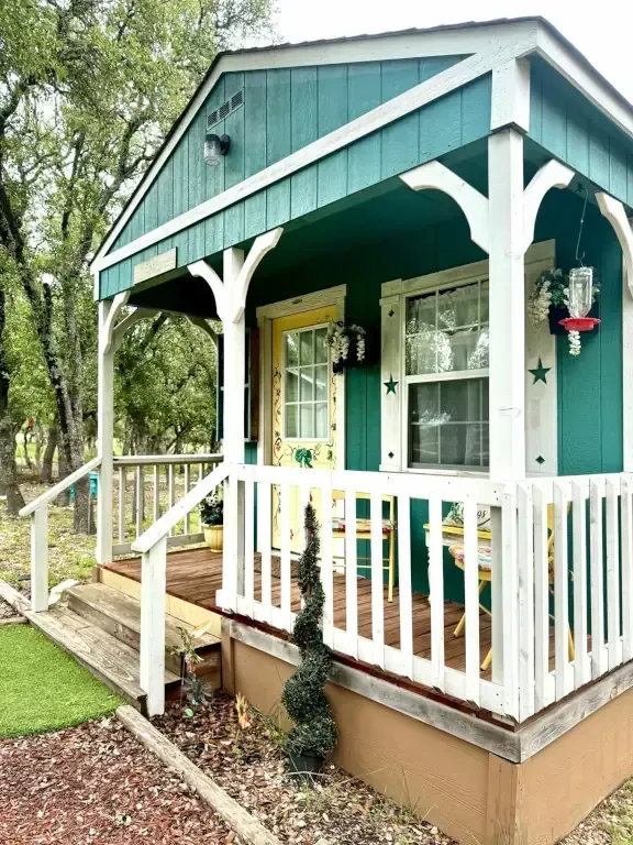 Small teal cabin porch with white railings, steps, and hanging flowers in a wooded setting