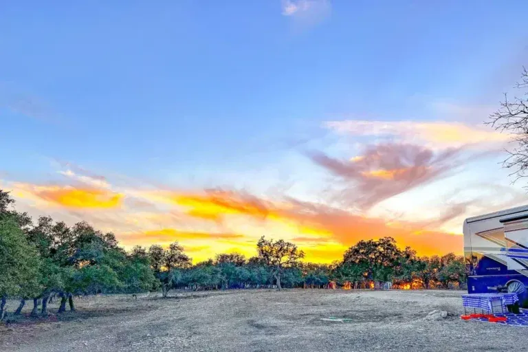 Sunset over a grassy field with trees and a blue car parked at the right edge