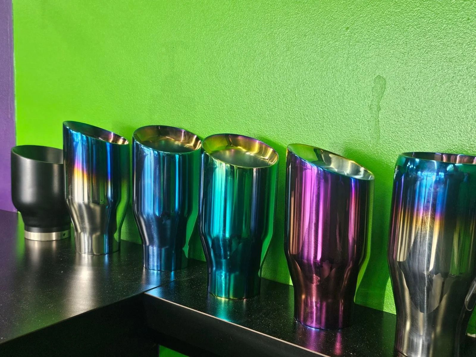 Five iridescent stainless steel tumblers on a black table against a bright green wall