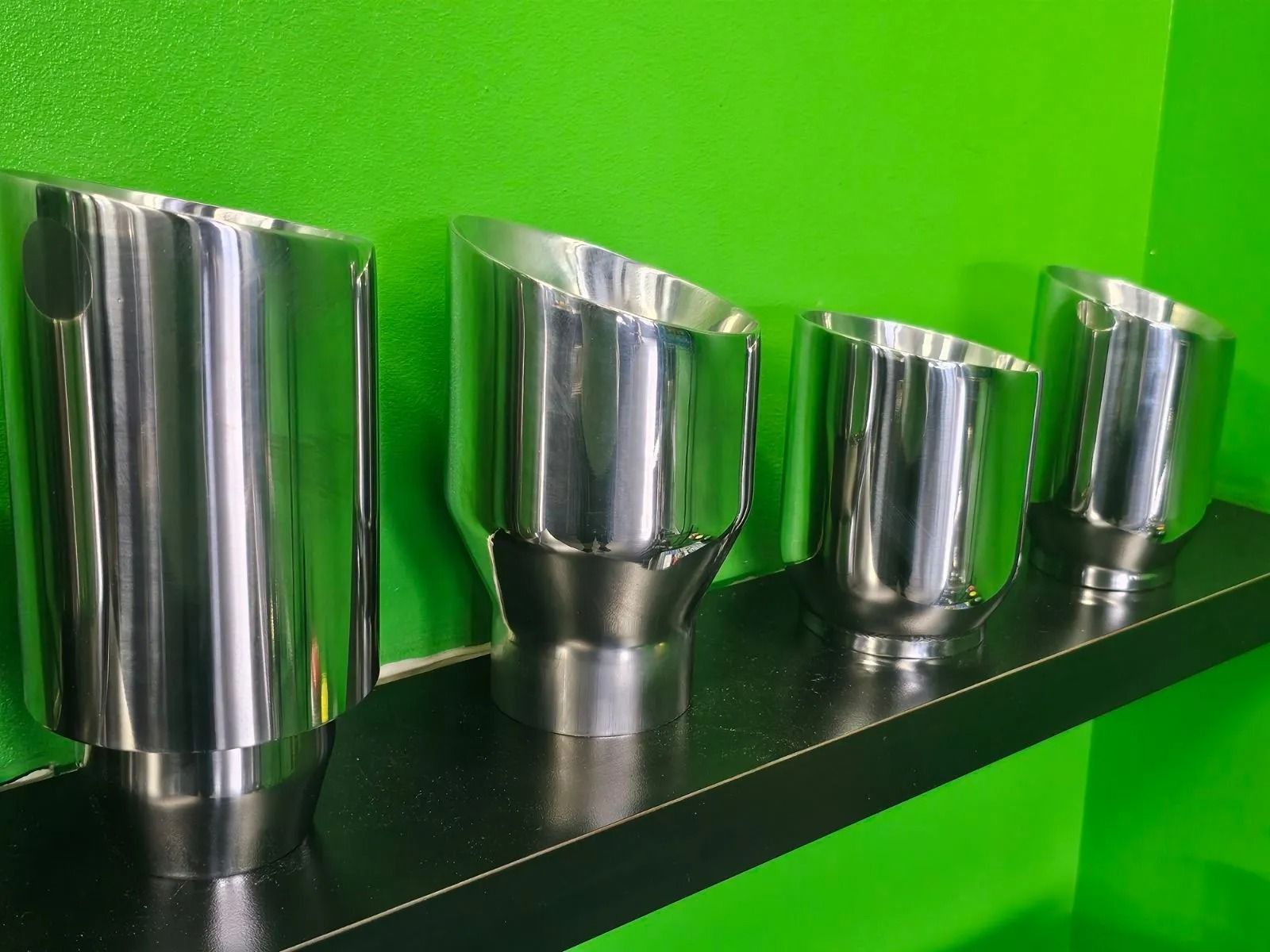 Four shiny metal cups displayed in a row on a black shelf against a bright green background