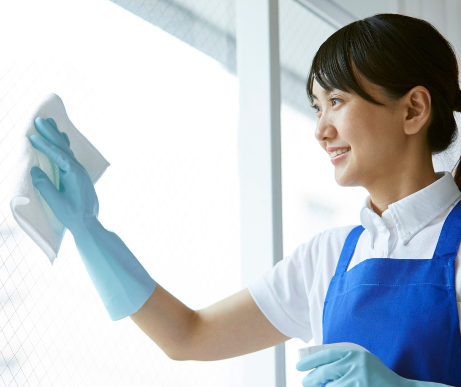A cleaner in blue apron and gloves wipes a bright window with a cloth, smiling.