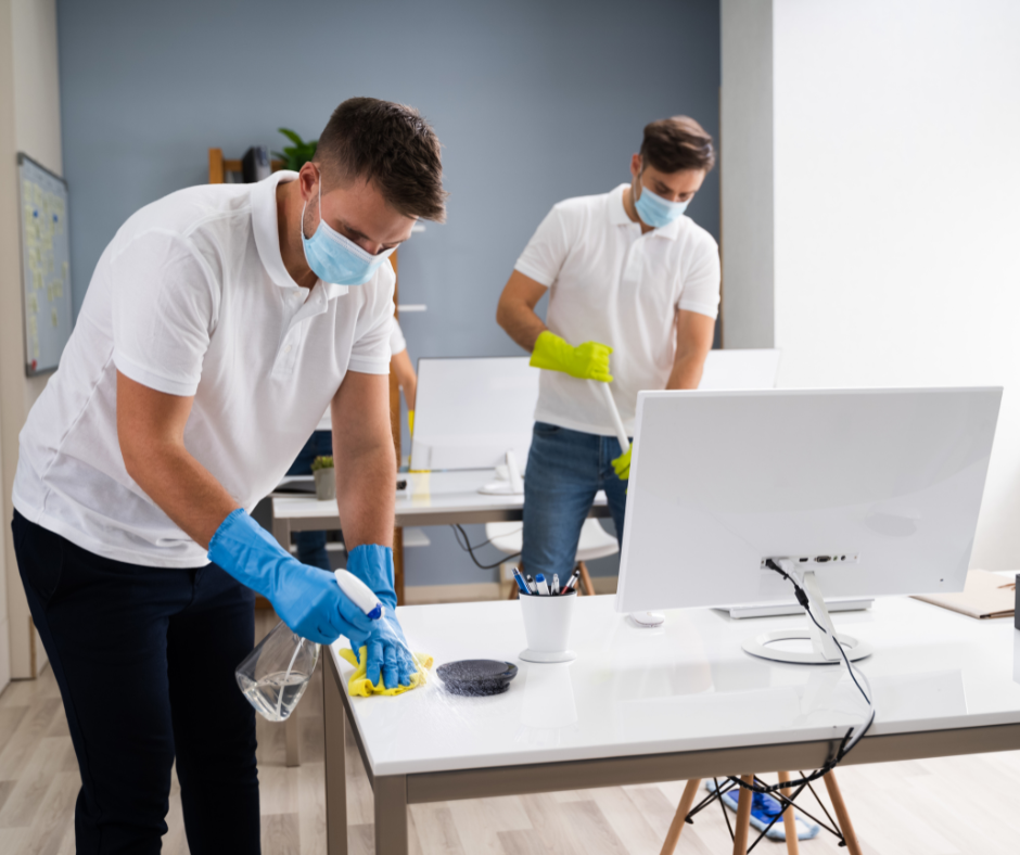 Two cleaners disinfecting a desk in an office, wearing gloves and masks