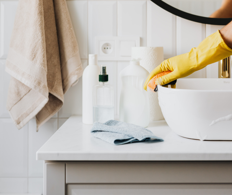 Gloved hand cleaning a bathroom sink with soap and towel on a white counter