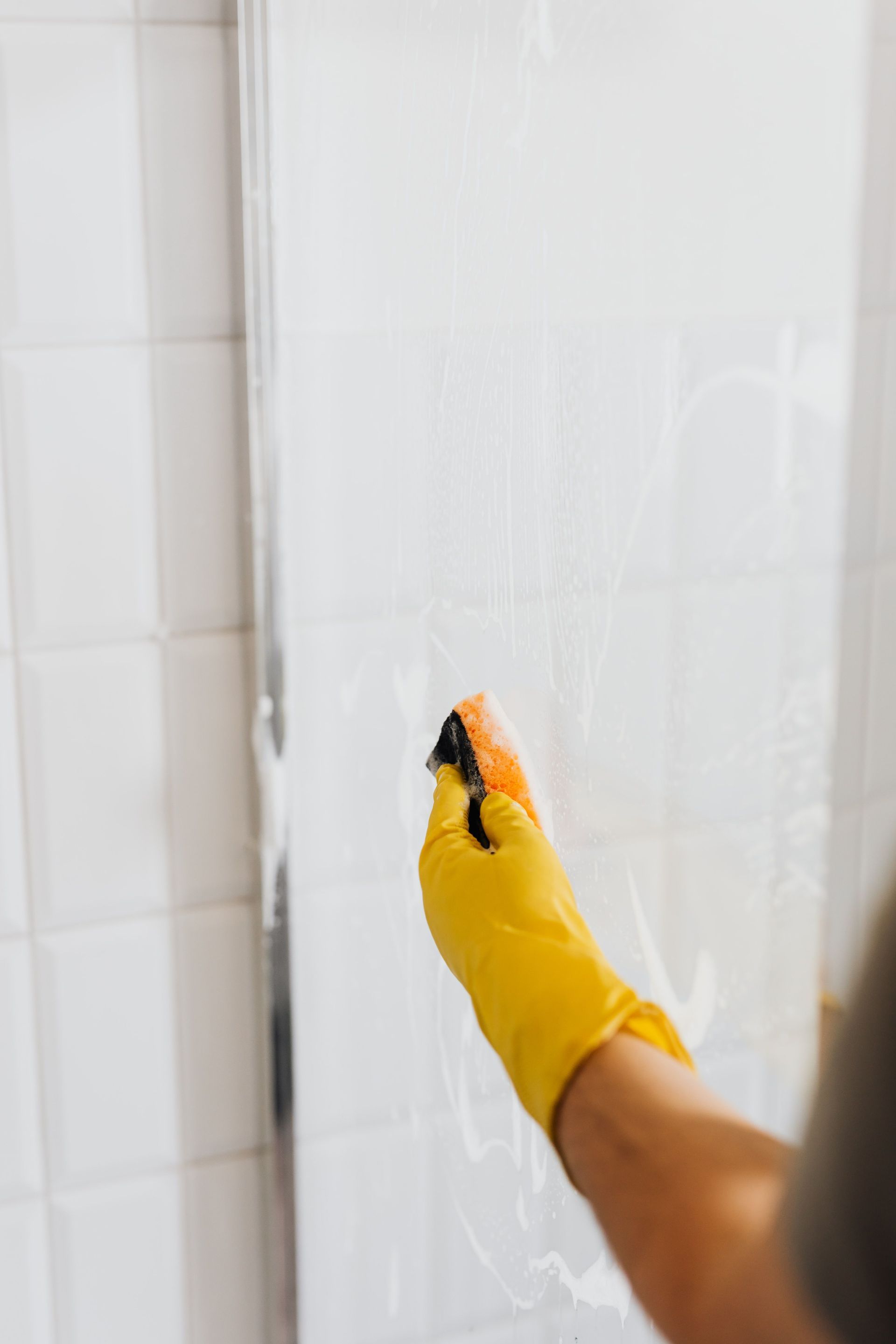 Gloved hand scrubbing a white tiled wall with cleaning spray.