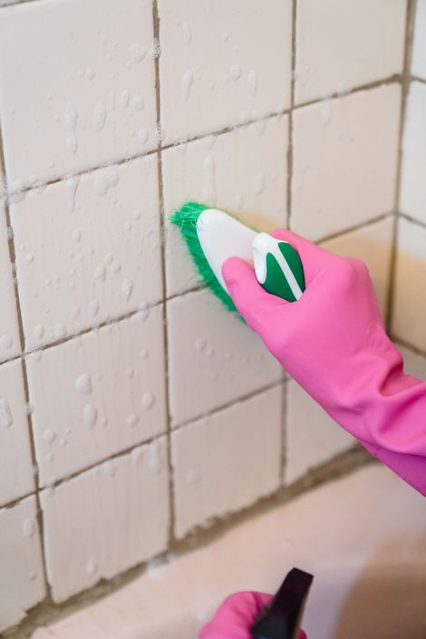 Pink-gloved hand scrubbing white bathroom tiles with a green sponge