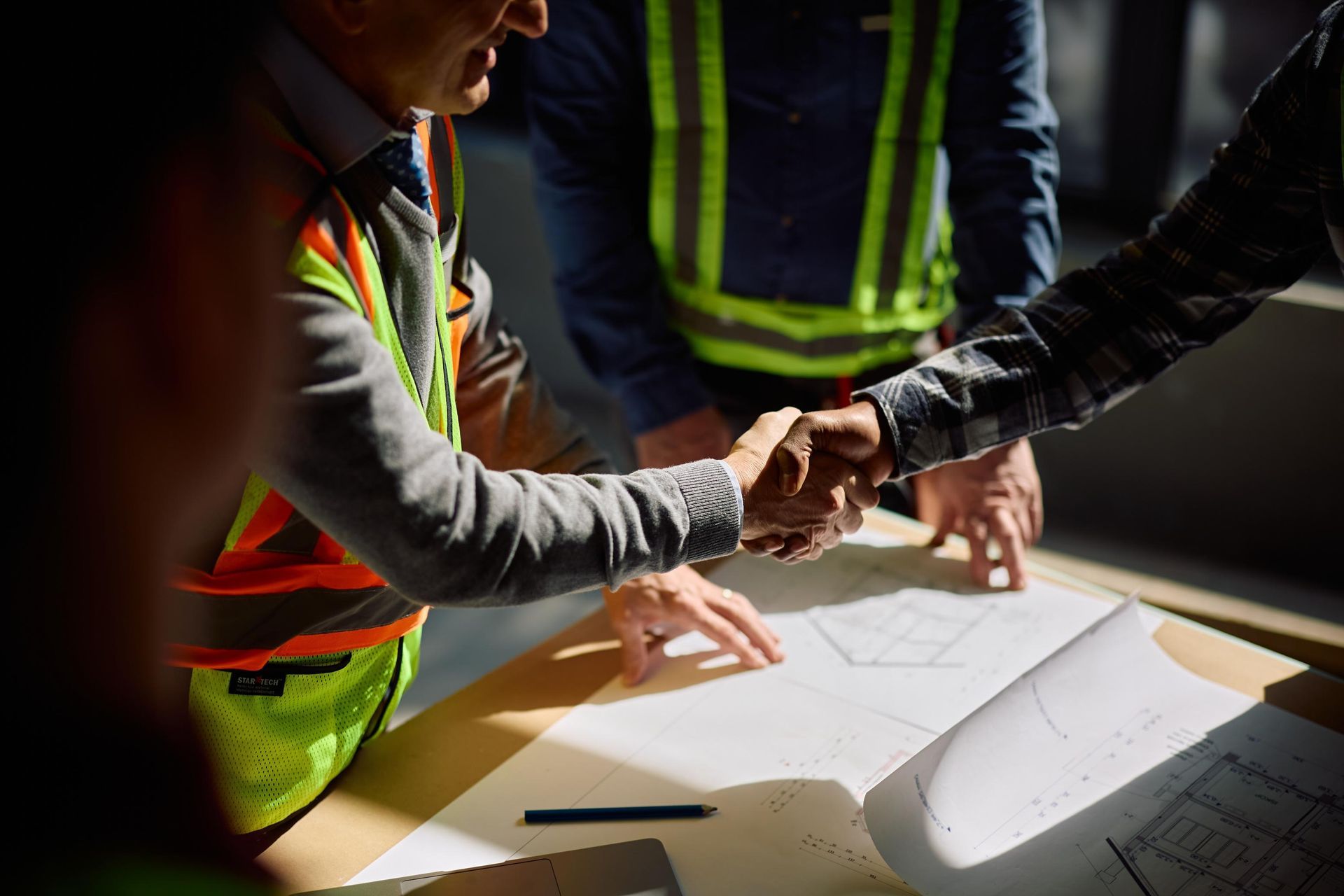 Workers in safety vests shake hands over a table with plans at a construction site