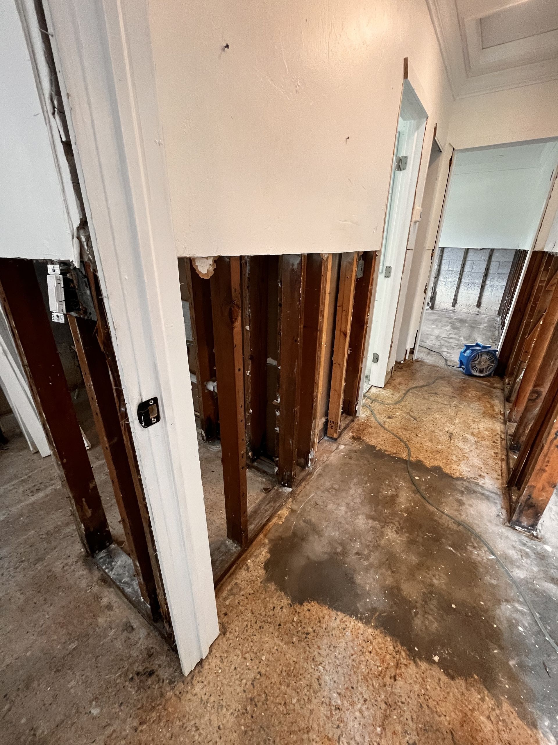 Hallway under construction, wall partially removed, revealing wooden studs. Concrete floor with debris.