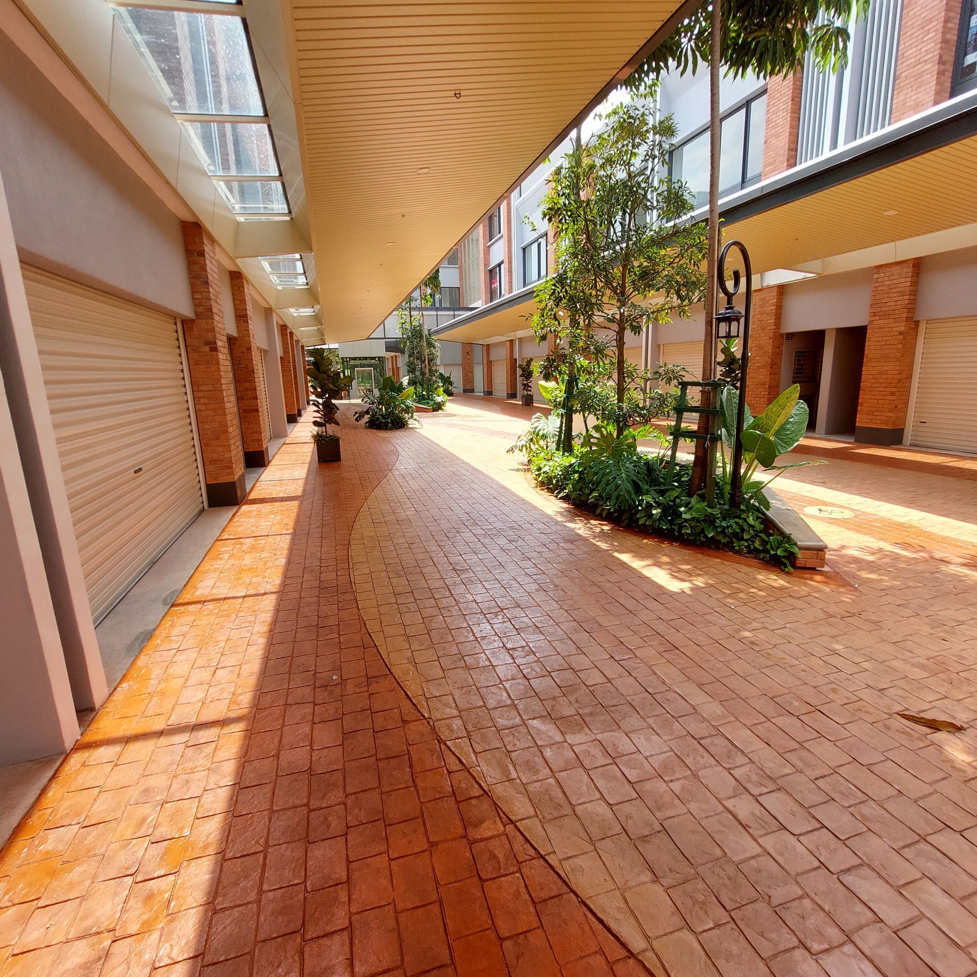 Brick-paved walkway lined with shops, awnings, and plants. Bright sunlight, neutral colors.