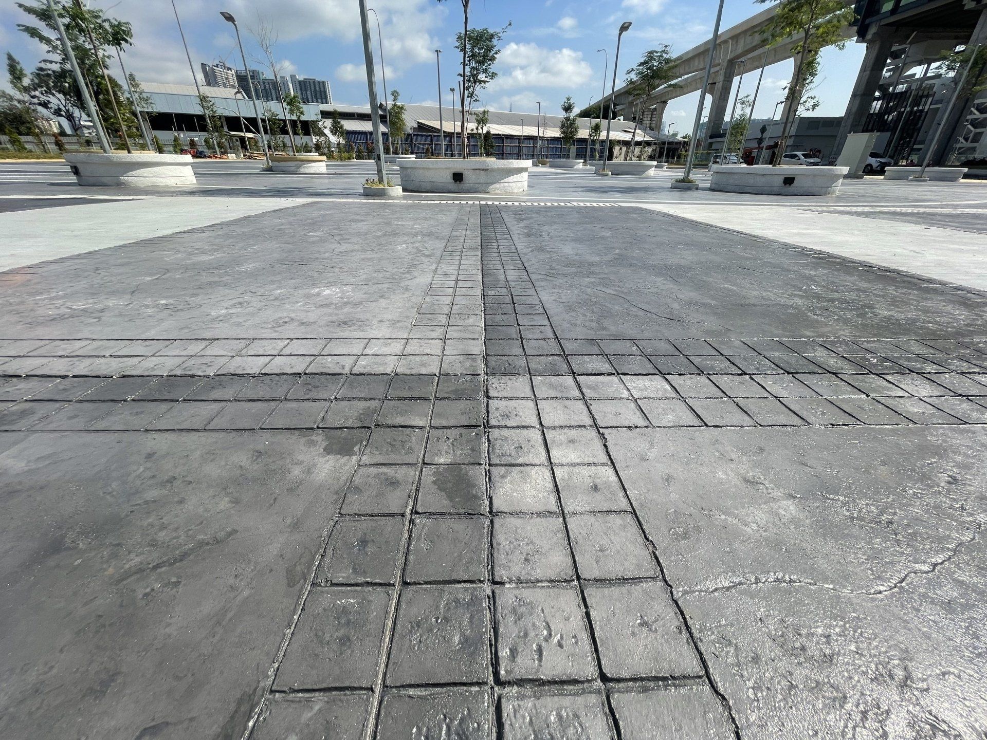 Gray concrete plaza with patterned pathways and surrounding structures under a cloudy sky.