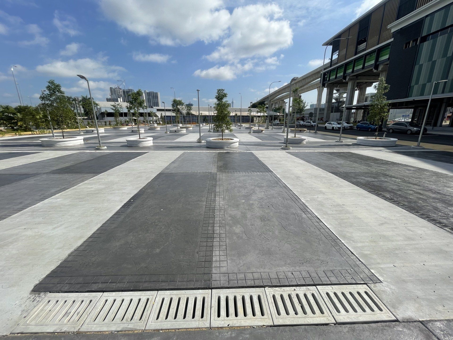Outdoor plaza with patterned pavement and drainage grates; train tracks and modern building in background.