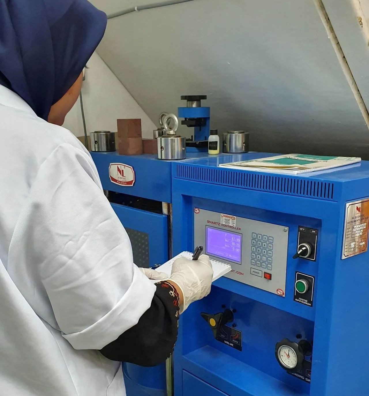 A person in a lab coat and hijab operates a blue testing machine, taking notes on a clipboard.