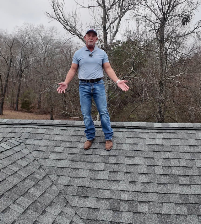 Man standing on a shingled roof with arms outstretched in front of leafless trees