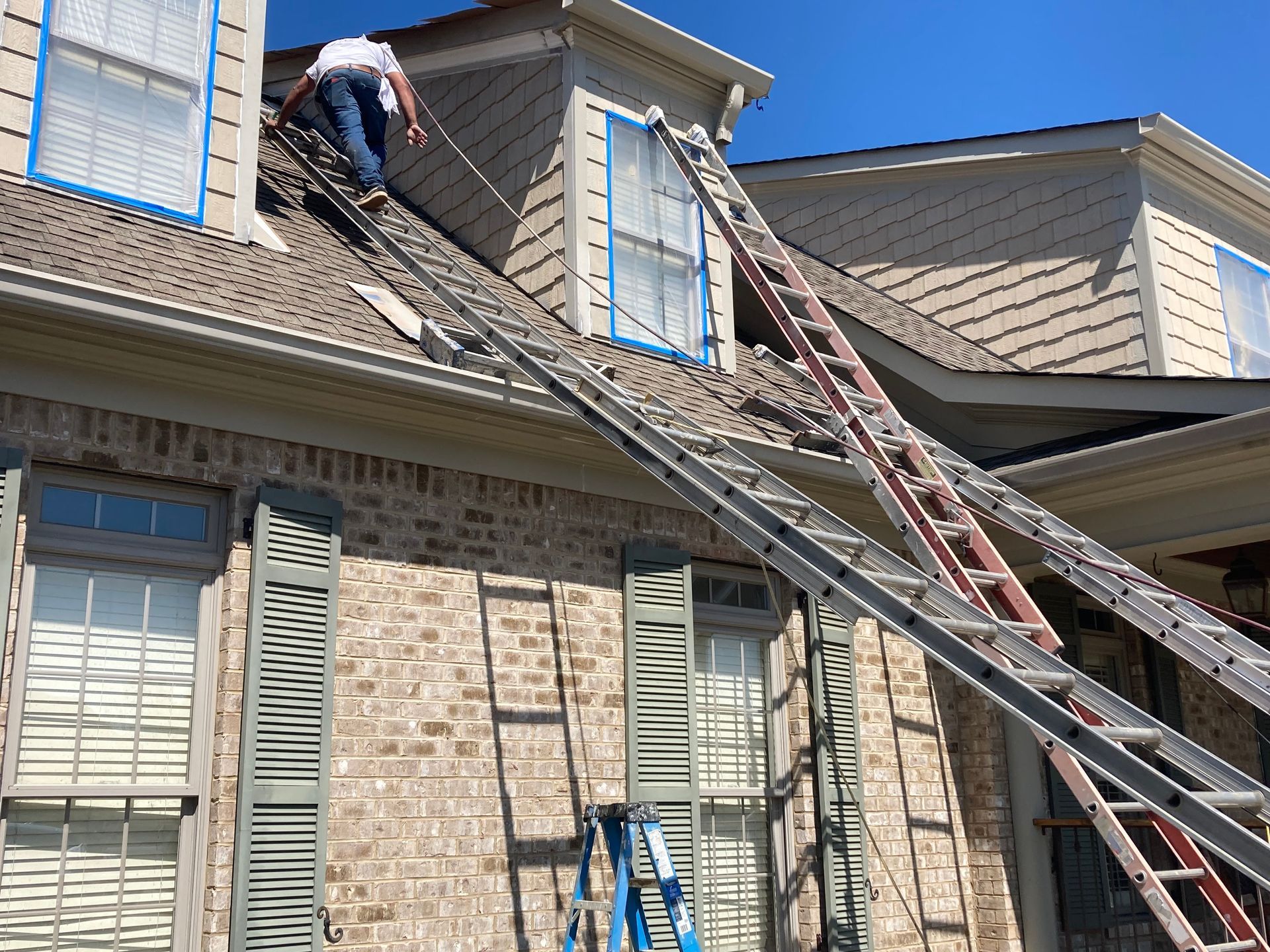 Worker on a ladder repairing a tan brick house roof beside a second ladder and blue tarp