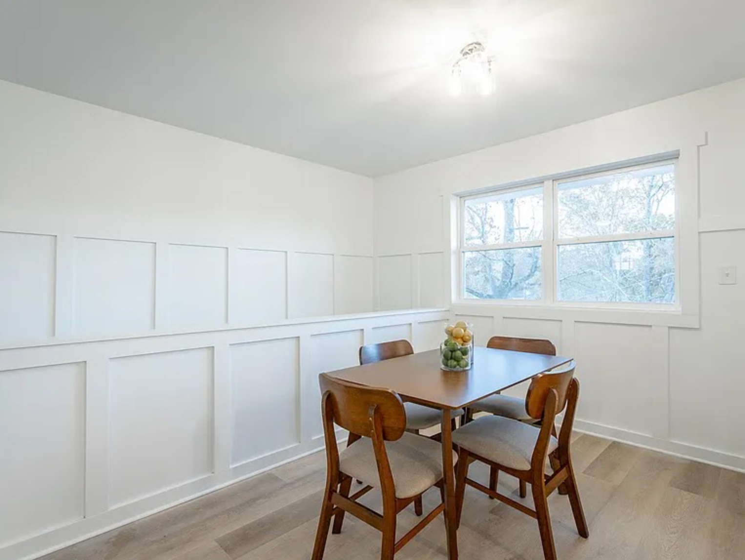 Bright dining room with white paneled walls, a wooden table, chairs, and a large window.