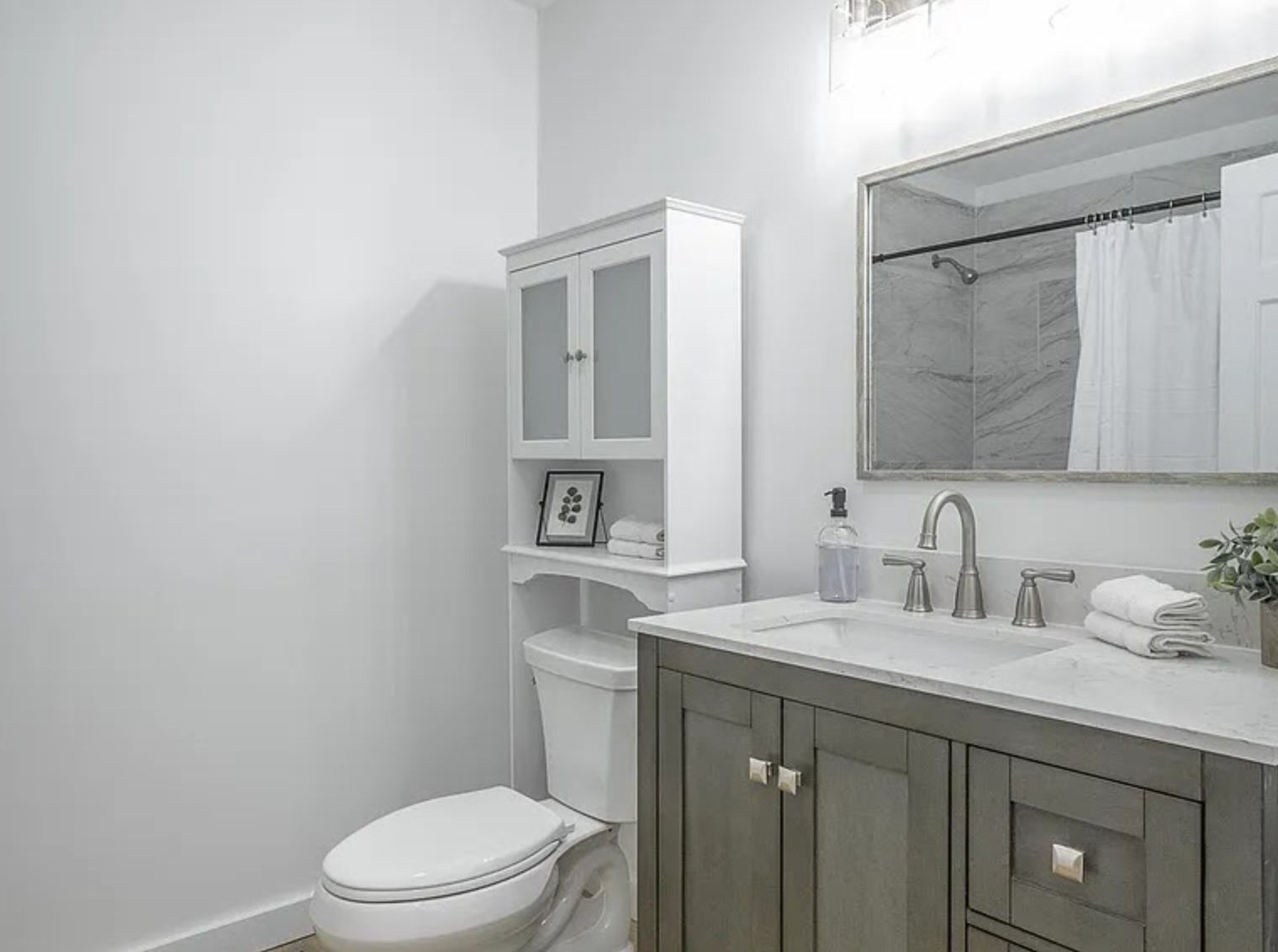 Bright white bathroom with gray vanity, toilet, mirror, and small cabinet.