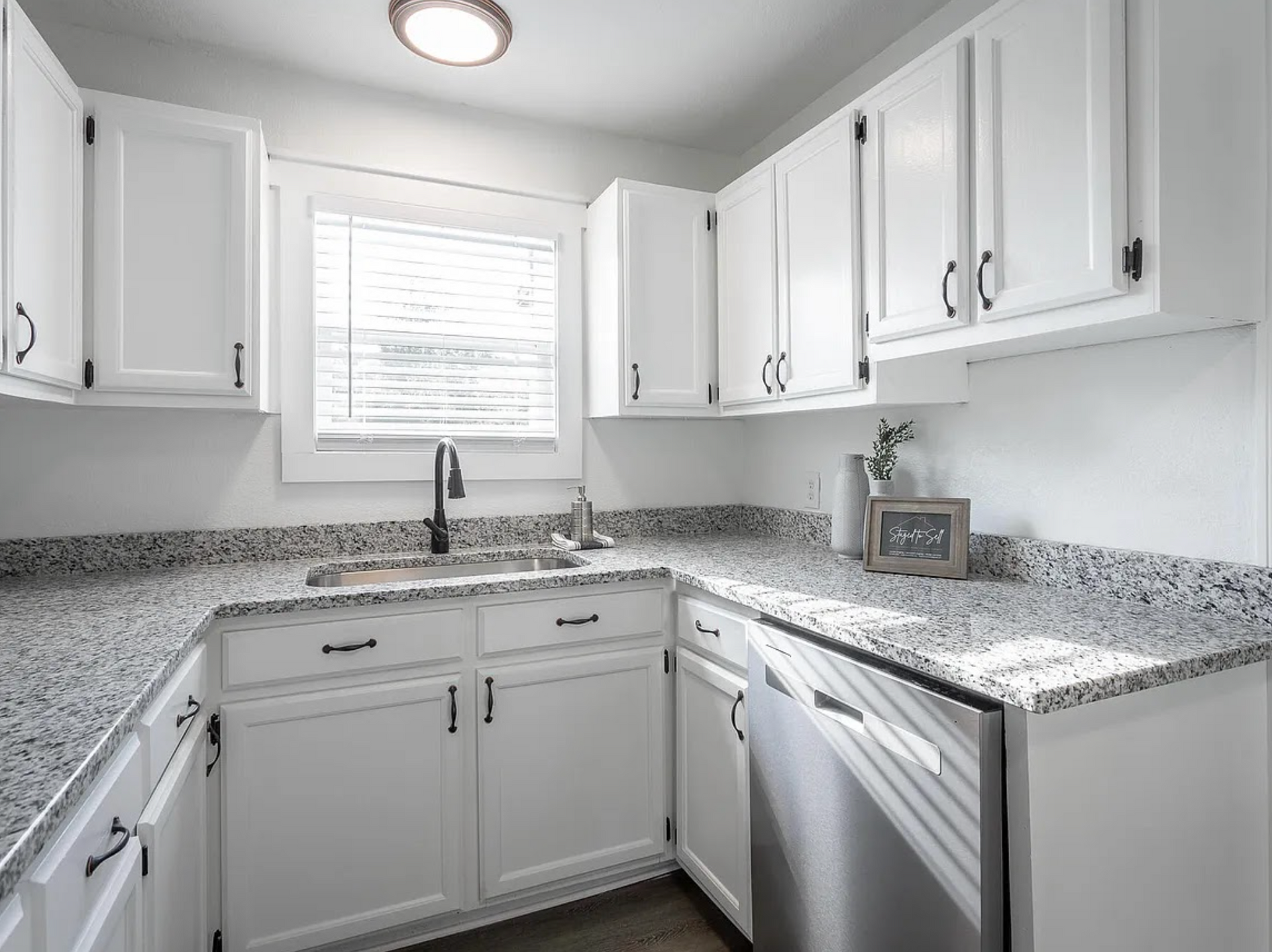 Bright white kitchen with granite countertops, white cabinets, sink under window, and dishwasher.