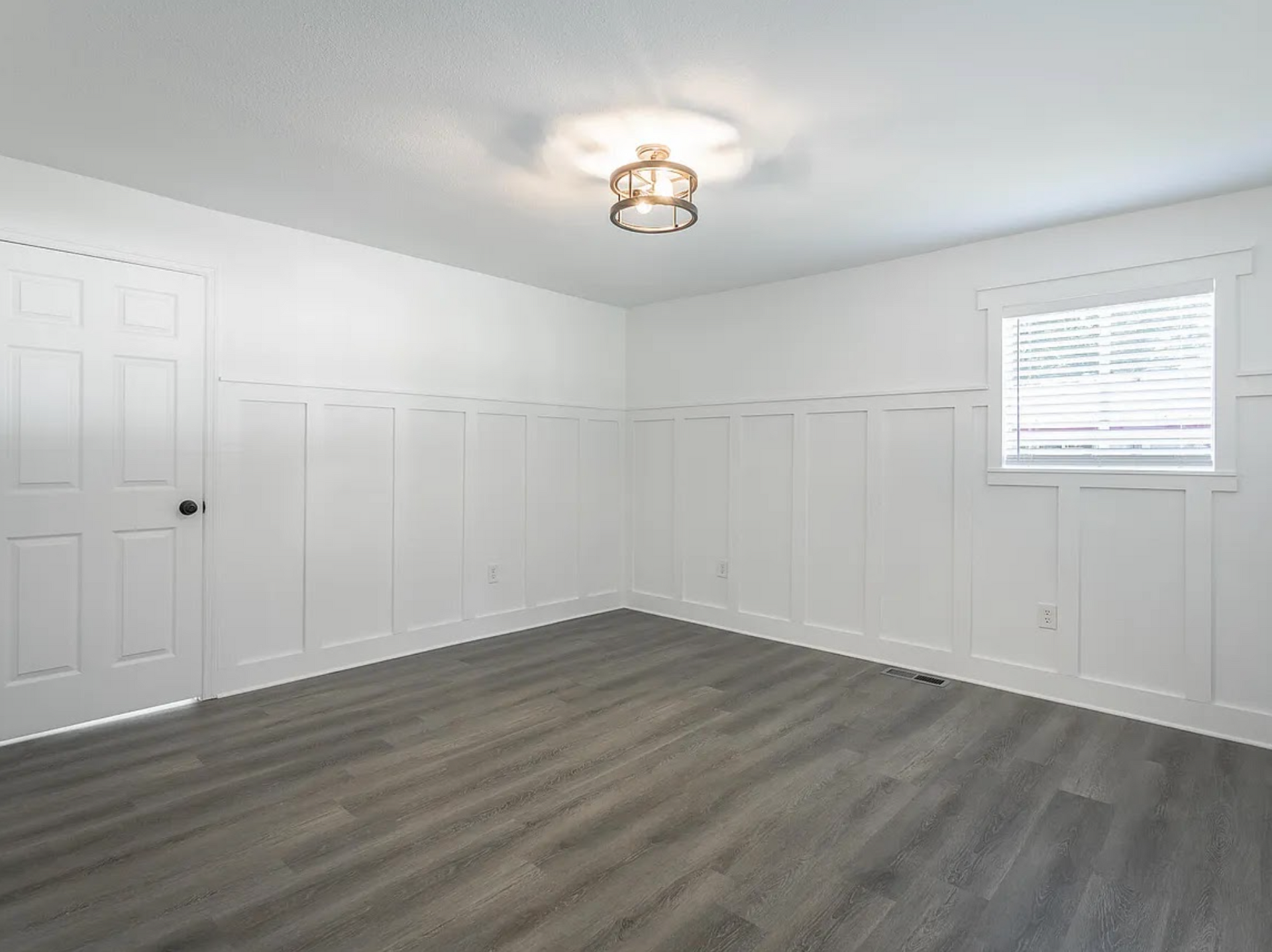 Empty white room with gray wood floors, paneled walls, a ceiling light, and a window with blinds