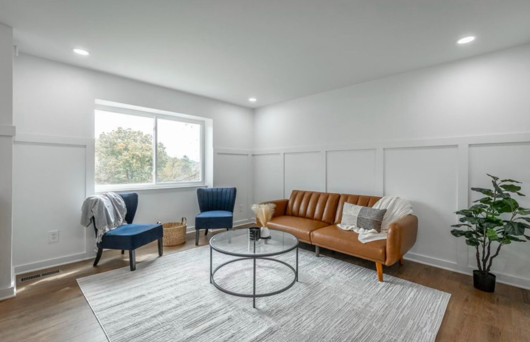Bright modern living room with a tan sofa, blue chairs, round coffee table, and large window.