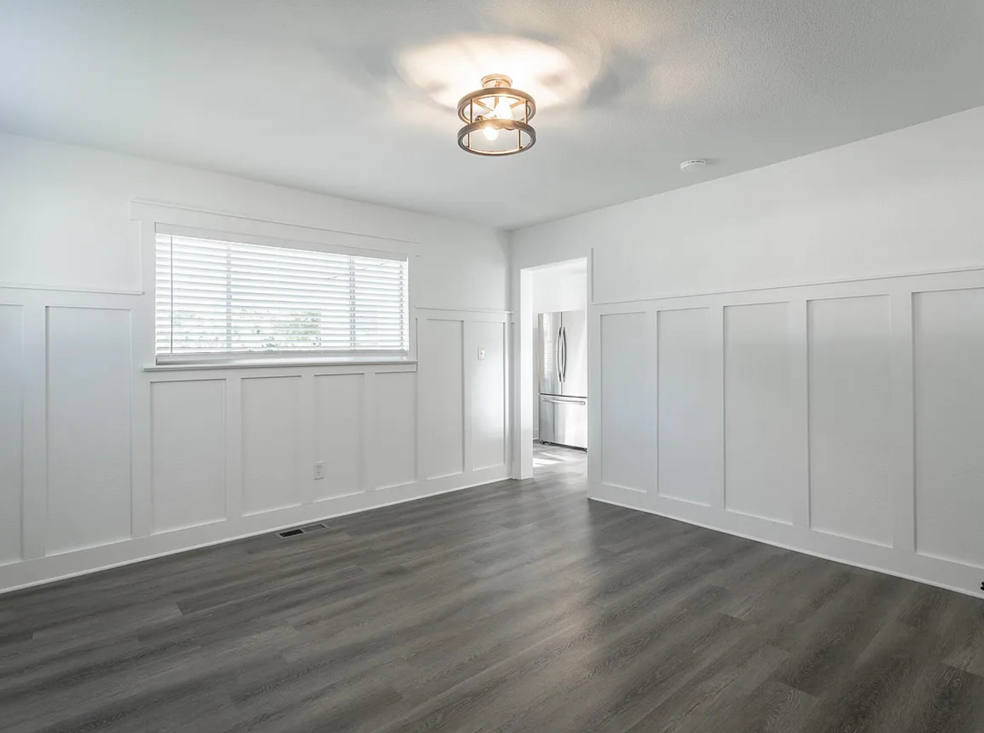 Empty white bedroom with gray wood flooring, window blinds, and paneled walls.