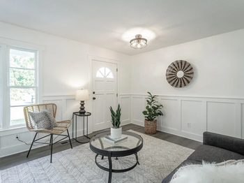 Bright white living room with gray rug, round coffee table, woven chair, plants, and wall decor