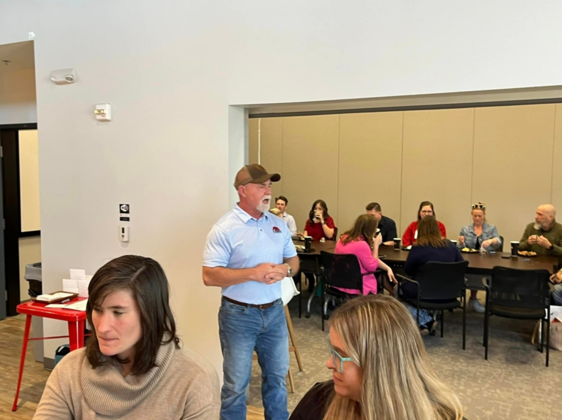 Man speaking to seated audience in a bright meeting room with tables and chairs.