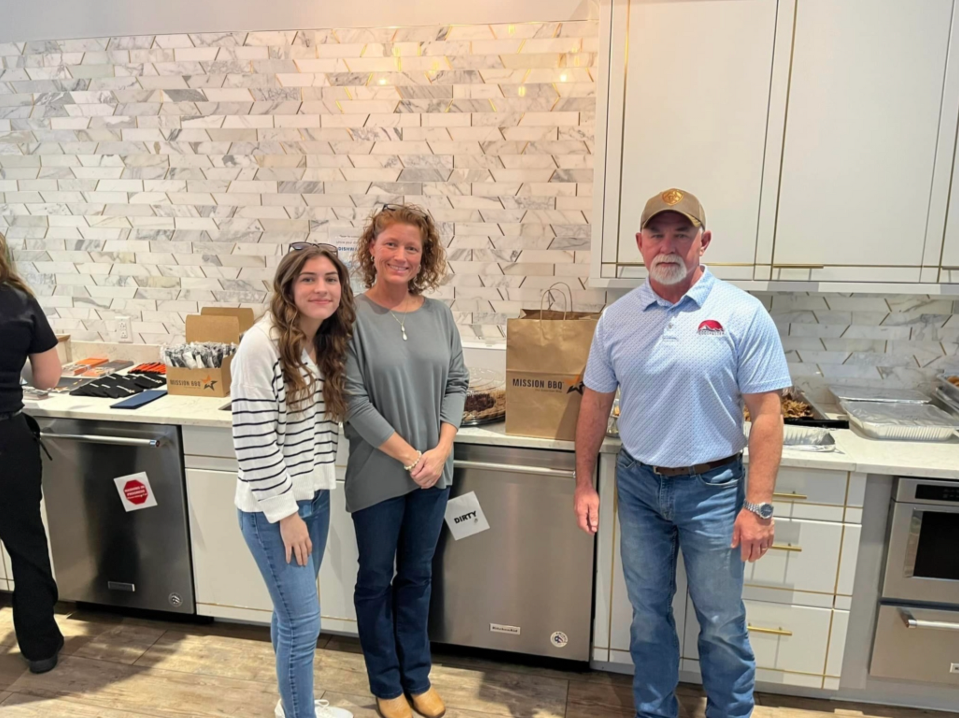 Three people standing in a kitchen beside stainless steel appliances and white cabinets