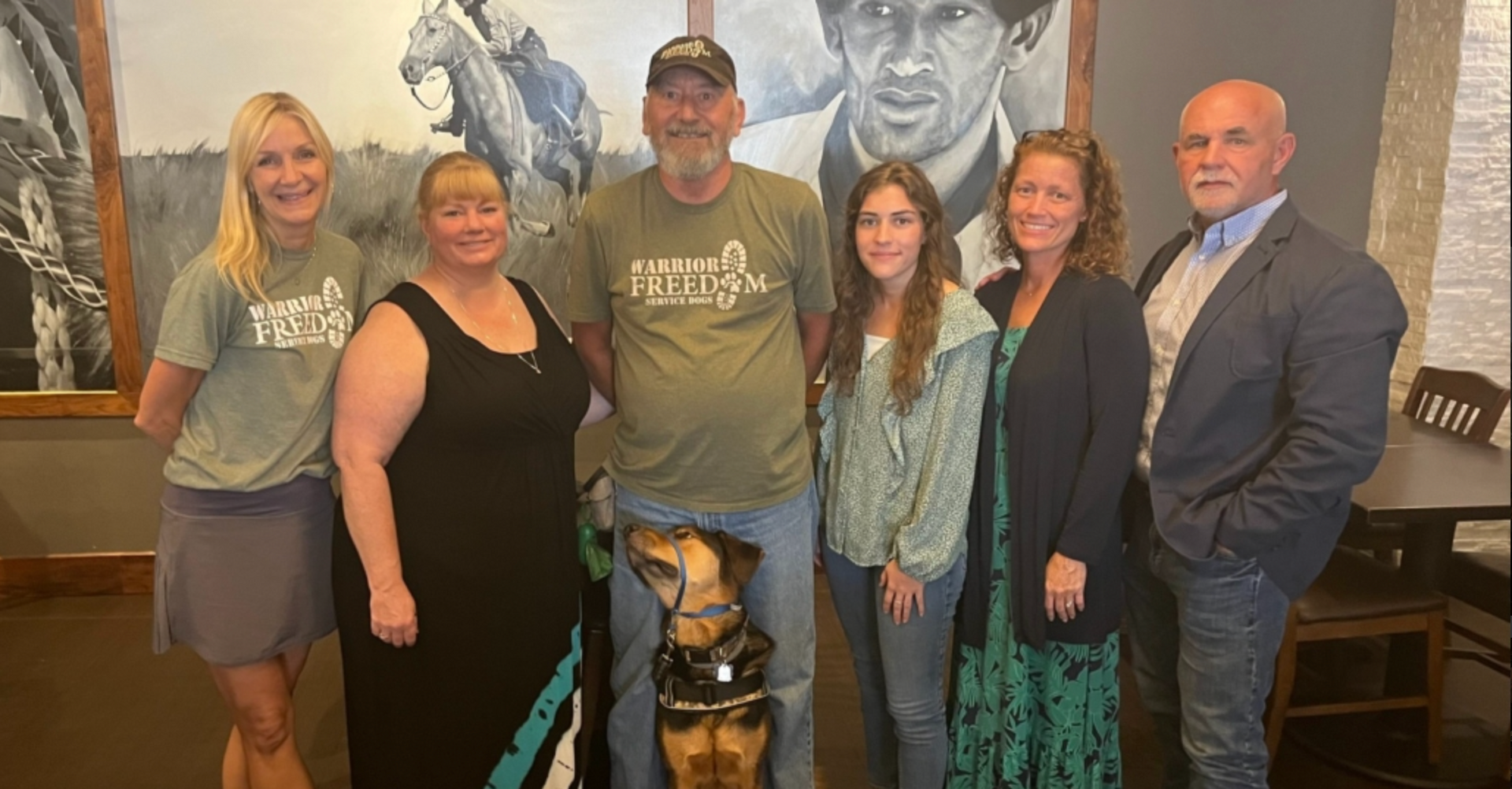 Group of six smiling people posing indoors in front of a mural, with a dog in the center.