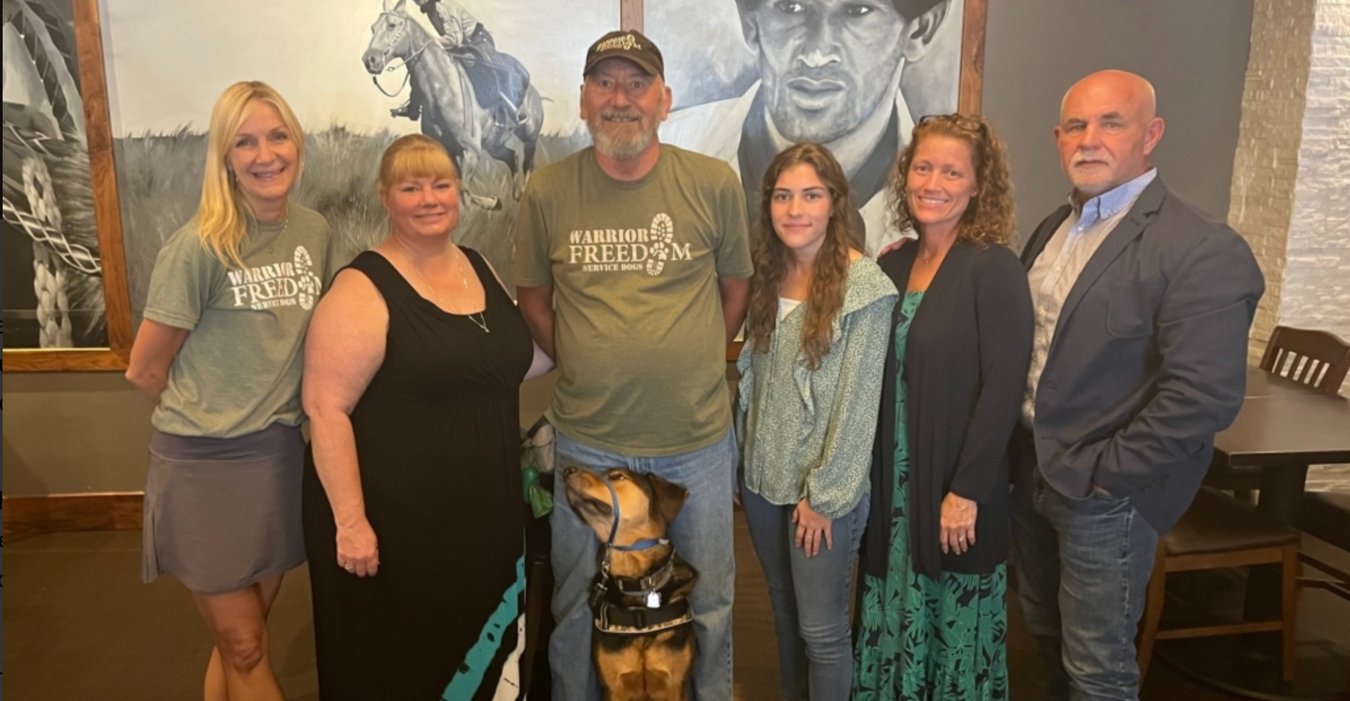 Six people pose indoors in front of a mural, with a service dog centered in front.