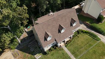 Aerial view of a brown-roofed house with dormer windows, surrounded by trees and a grassy yard.