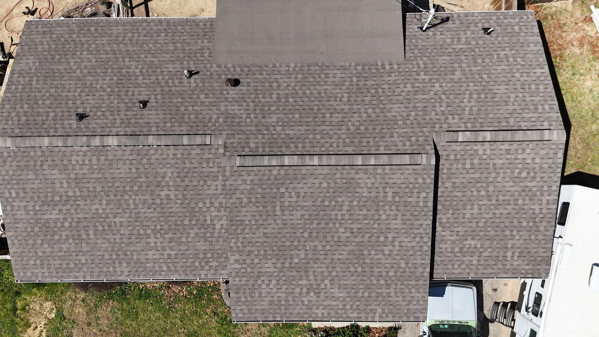 Aerial view of a gray shingle roof with multiple sections and vents beside a driveway and greenery