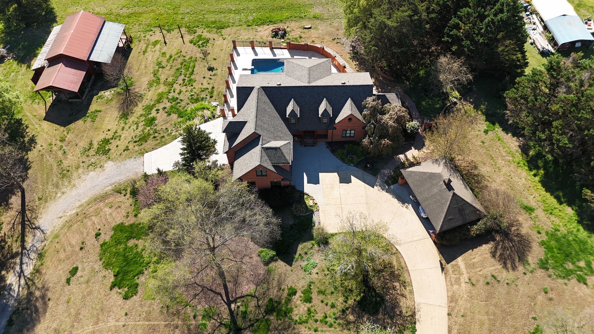 Aerial view of a rural house with red roofs, a driveway, and surrounding trees and grassy land.